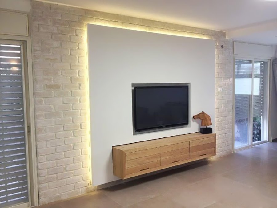 Parete in cartongesso elegante e semplice illuminata sui bordi che proietta una bellissima luce sul muro in mattoni a vista verniciato in bianco. La struttura ospita la tv ed un modulo basso in legno.