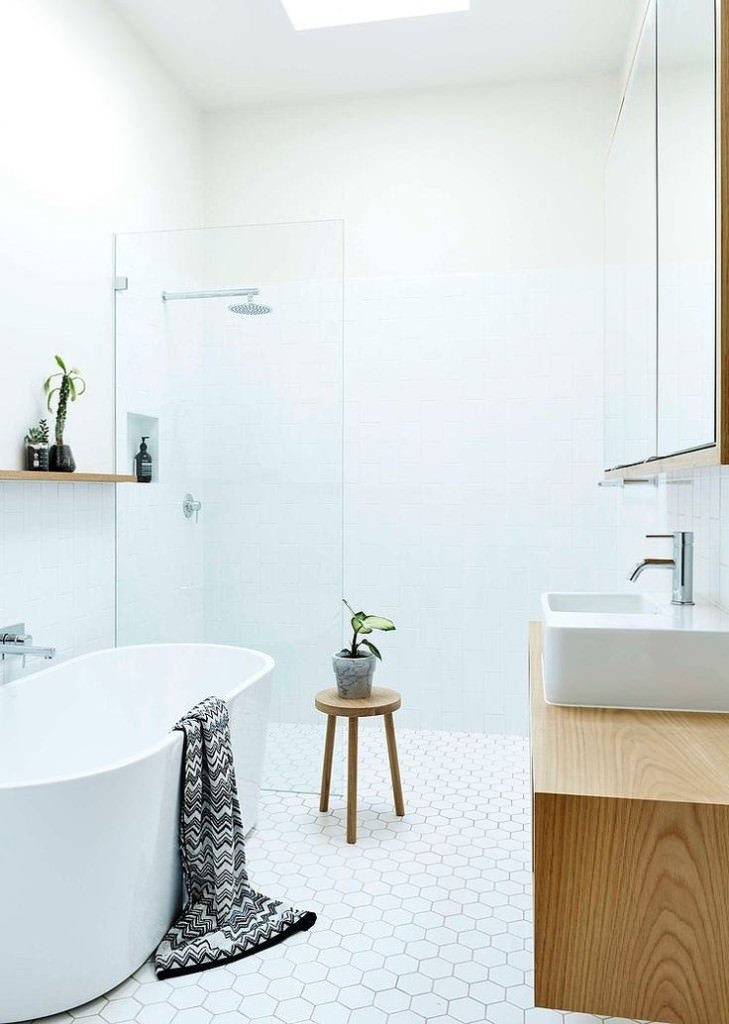 Un bagno totalmente bianco, con piastrelle esagonali sul pavimento e quadrate sulle pareti. Sono stati inseriti un mobile sospeso e una mensola sopra la vasca in legno chiaro per rompere il total white dell'ambiente