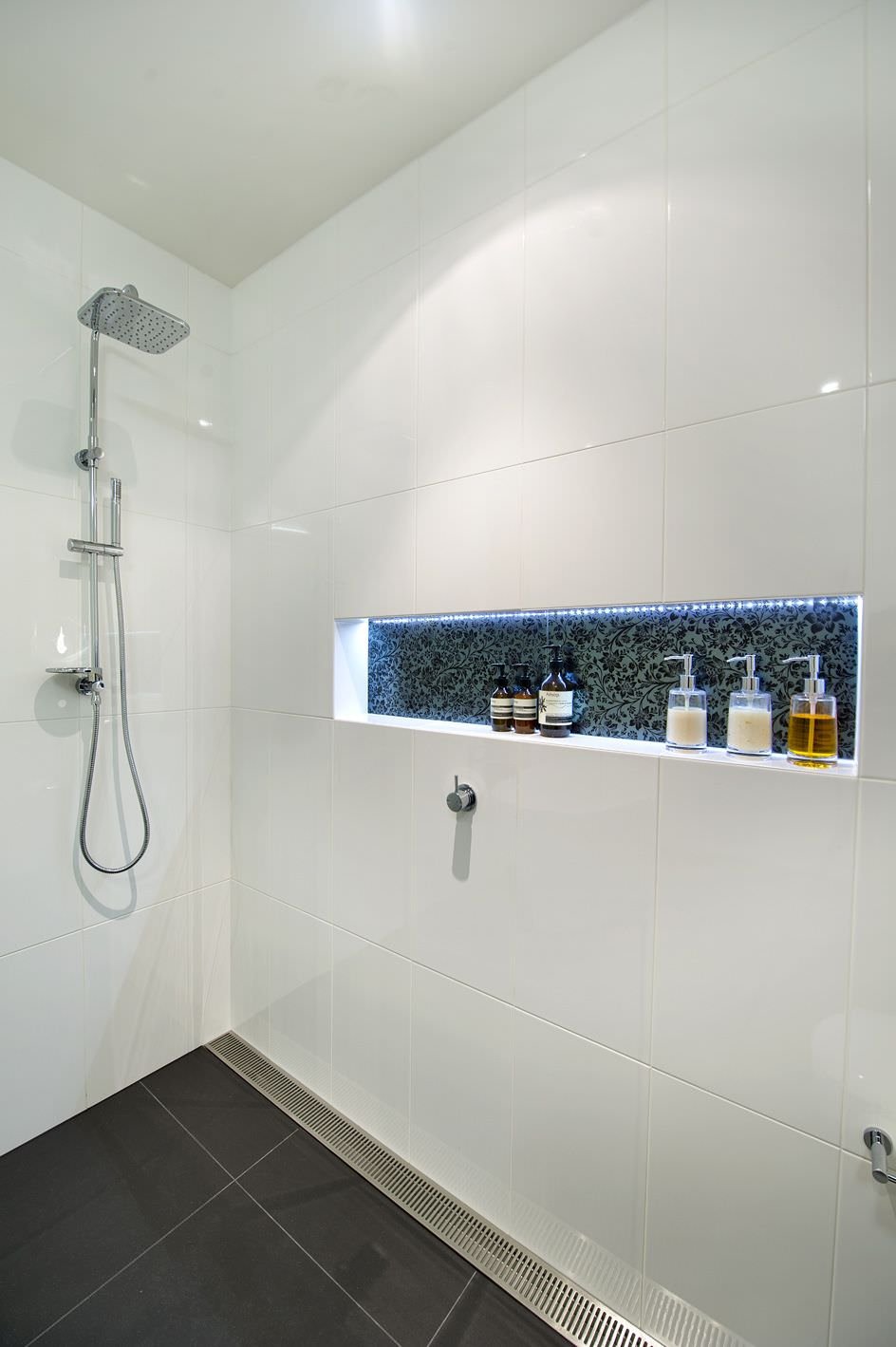 Idea per ristrutturazione bagno moderno con nicchia creata sulla parete della doccia. Rivestimento in ceramica bianca di grandi dimensioni, nicchia illuminata creata con piastrelle motivo floreale.