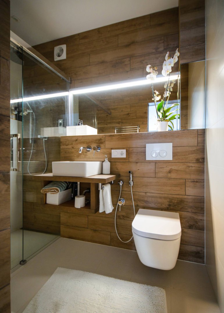 Progetto piccolo bagno con il rivestimento in listoni di gres porcellanato finto legno. Interessante l'idea della striscia di luce a led posizionata lungo lo specchio che allarga visivamente la stanza. Supporto lavabo in legno e wc sospeso