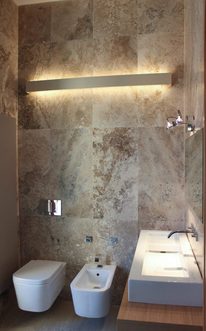 Ispirazione ristrutturazione piccolo bagno, moderno e particolare, con rivestimento in lastre di marmo, che aggiungono carattere alla stanza. Una lunga lampada, posizionata sul muro assicura la giusta illuminazione. Sanitari sospesi ed un lungo lavabo in ceramica per uno stile decisamente maschile. Idee bagni piccoli.