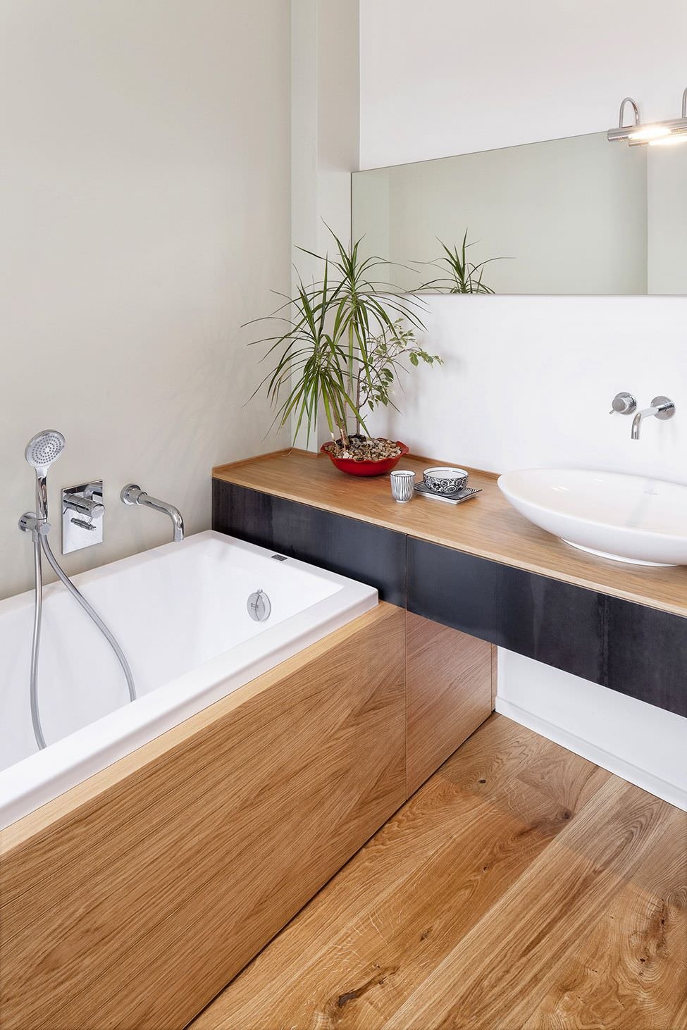 Immagine stanza da bagno di dimensioni ridotte con vasca. Il legno dei pavimenti, del top del mobile su cui appoggia il lavabo e del rivestimento vasca viene abbinato ai colori bianco e nero.