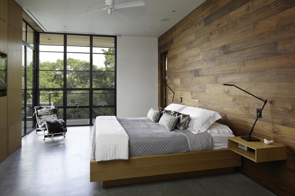 Modern bedroom with the wall along the headboard covered in wood.  Minimal style with concrete floors and large windows with black profiles