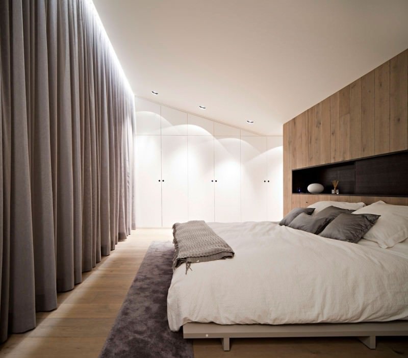 Modern bedroom with the headboard wall covered in wood (the same as the floor) which houses a niche with a decisive role in the design of the room.  Interesting light diffused along the windows and the ascending ceiling.