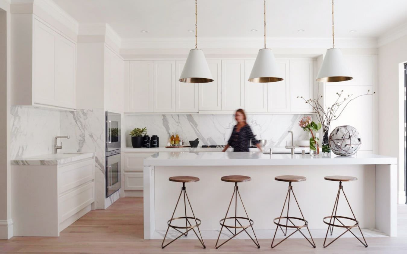 Cucina bianca con isola, stile contemporaneo, con mobili in laminato e top in marmo. Pavimento in legno e alcuni dettagli dorati (sgabelli e interno lampadari) 