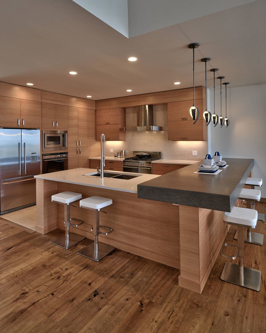 Cucina in legno con isola e penisola ad L con bancone bar. Top in cemento molto robusto che aggiunge contrasto alla composizione. Controsoffitto in cartongesso con faretti da incasso e lampade a sospensione.