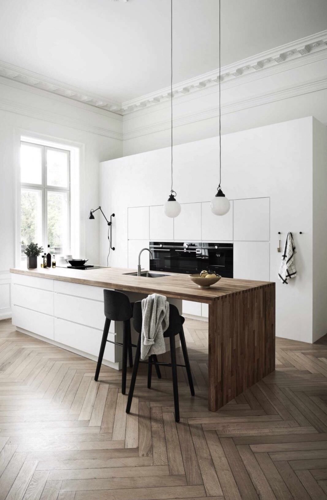 Cucina con isola moderna, stile parigino, in laminato bianco ed il top, con piano di cottura a induzione, realizzato in legno. Parete attrezzata a filo muro. Pavimenti in legno a spina di pesce e sgabelli neri che aggiungono contrasto e dinamicità.