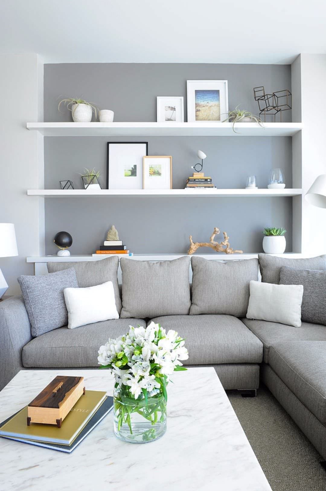 Stupendo soggiorno scandinavo moderno ed elegante, con il dettaglio di una scaffalatura creata all'interno di una nicchia del muro. Sopra le mensole sono posizionati piante, quadri e arte astratta. Pareti in toni di grigio, come il divano in stoffa.
