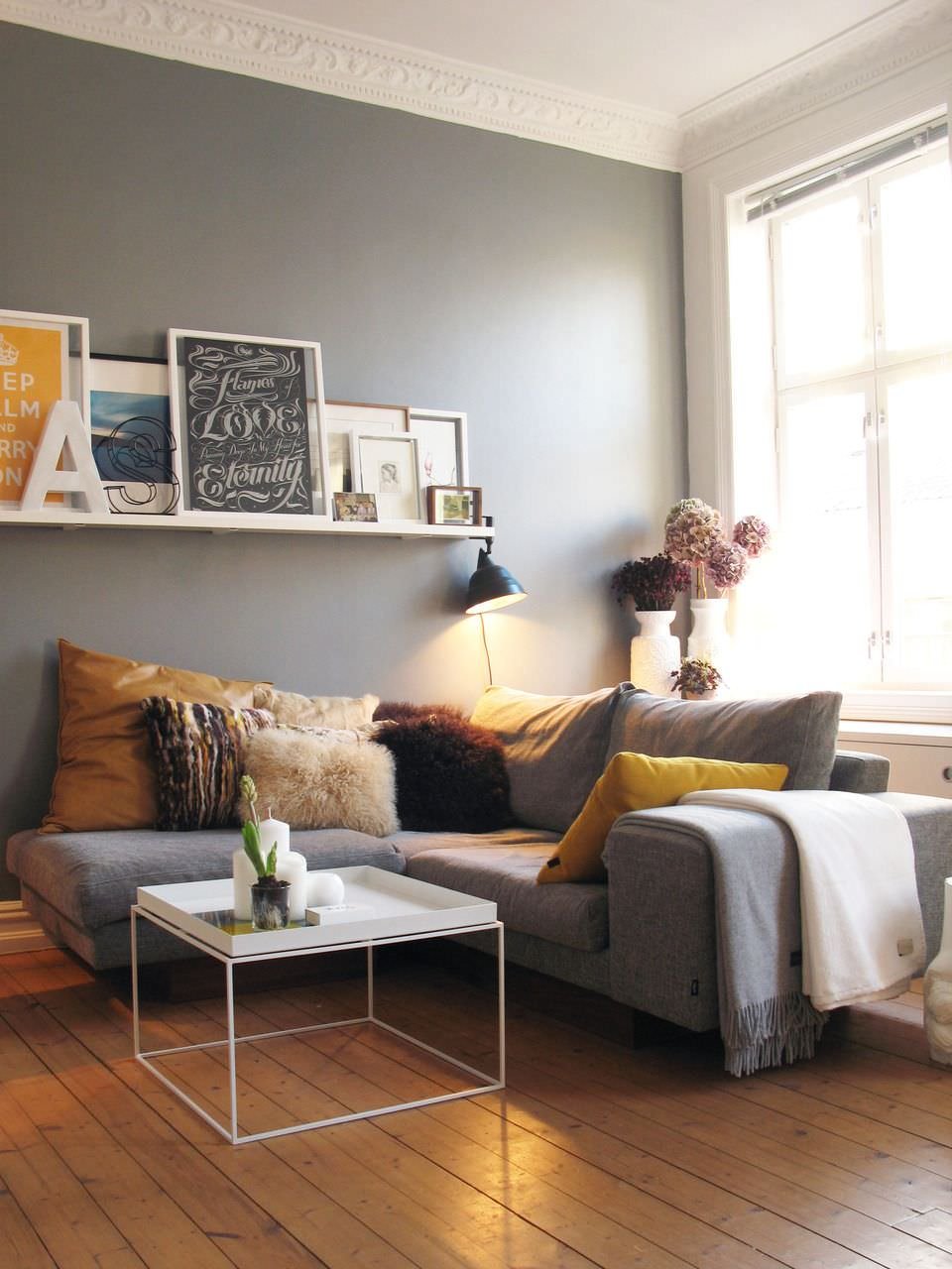 Bellissimo soggiorno scandinavo in tonalità più scure dello classico stile nordico: parete d'accento grigio e pavimento in teak marrone caldo. Cuscino e alcune decorazioni in tonalità di giallo e marrone in linea con il parquet.