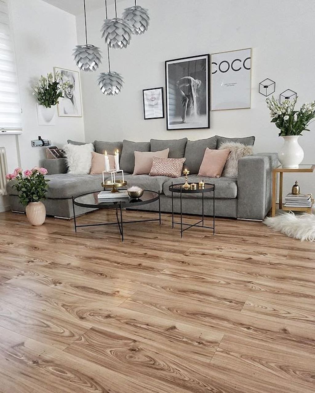 Salotto scandinavo femminile ricco di elementi. Divano grigio ad L in stoffa con cuscini rosa spento. Alcuni dettagli metallici: oro e argento. Parquet in legno.
