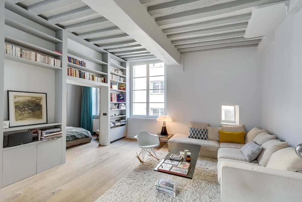 Living scandinavo contemporaneo con le pareti, la libreria in cartongesso e le travi a vista tinteggiate in grigio chiaro. Pavimento in rovere. I libri ed alcuni cuscini offrono il giusto tocco di colore.
