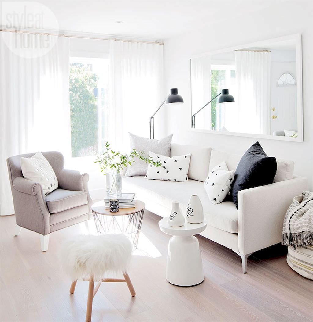 Salotto nordico di piccole dimensioni, allargato visivamente dalla luce riflessa dalle pareti bianche e dallo specchio usato come un quadro. Pavimento in legno chiaro, divani in tessuto bianco e poltrona beige. Alcuni contrasti di nero.